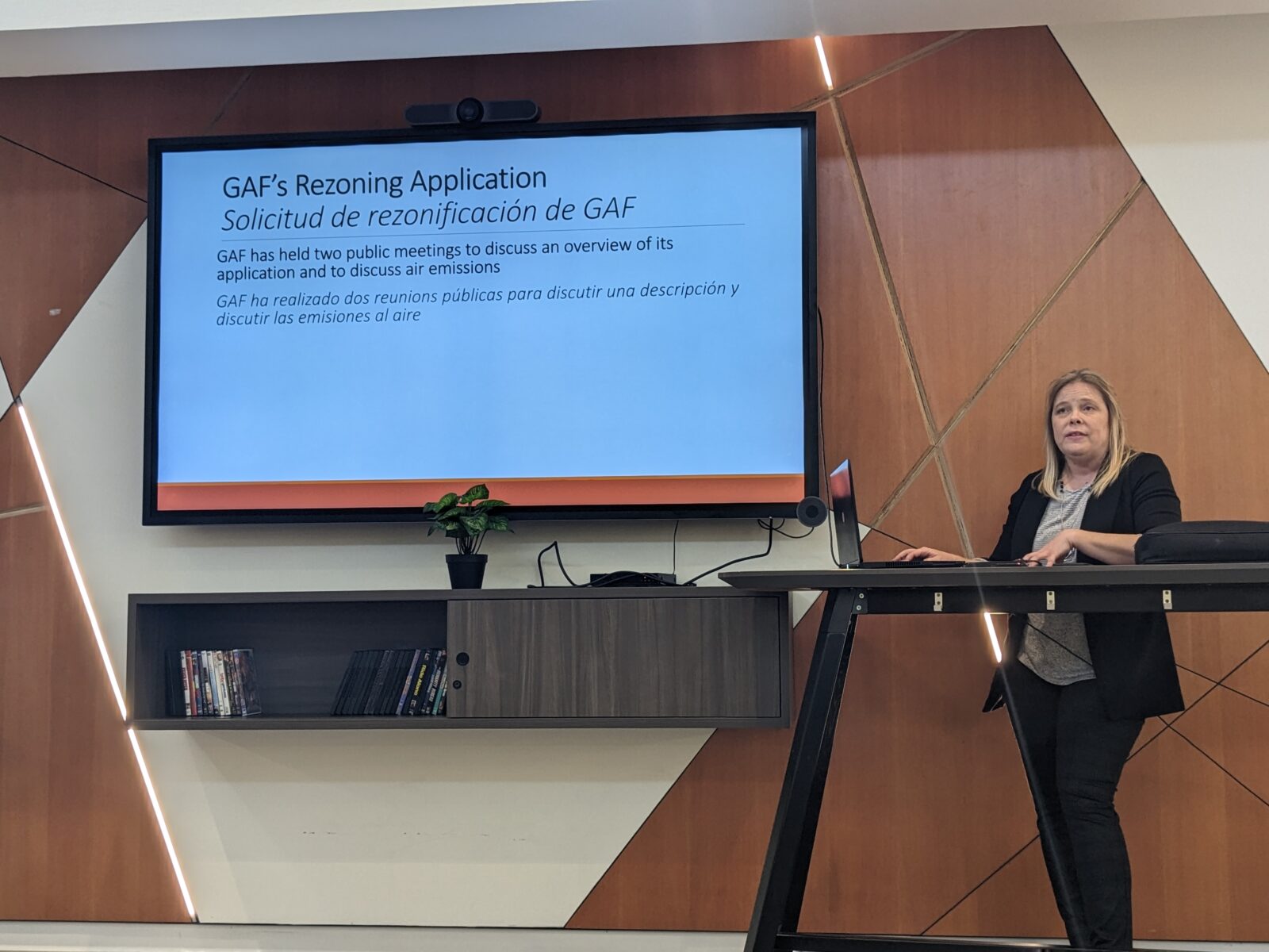 Wendi Hammond, an attorney with Legal Aid of Northwest Texas, stands next to a screen displaying a presentation about environmental justice concerns in West Dallas. The screen reads: "GAF's Rezoning Application: GAF has held two public meetings to discuss an overview of its application and to discuss air emissions."
